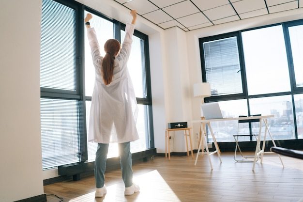 back-view-female-doctor-white-coat-stretching-hands-background-window-sunny-day_482921-256-6983684