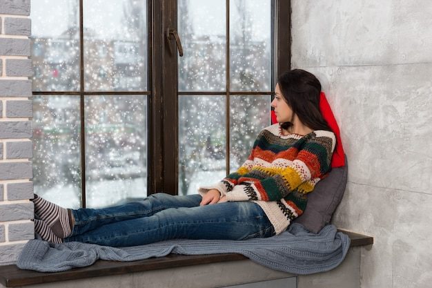 thoughtful-young-woman-warm-knitted-sweater-lying-down-pillows-windowsill-looking-out-window-while-snowing_385506-5794-8101529